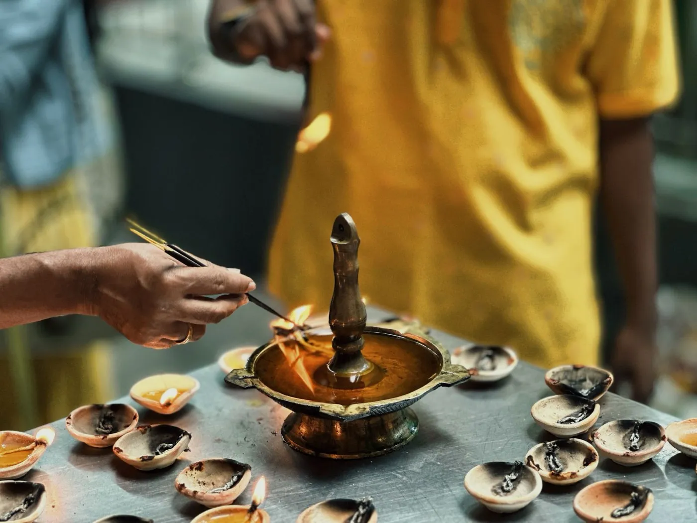 Hands lighting a brass oil lamp surrounded by rows of small clay lamps as an offering