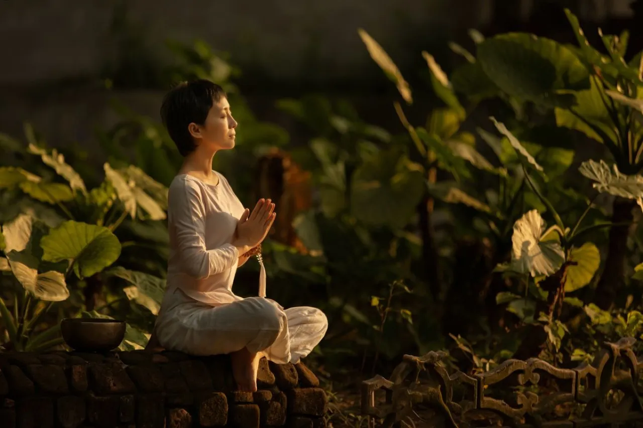 A person in white meditating at golden hour beside a singing bowl and prayer beads among lotus leaves