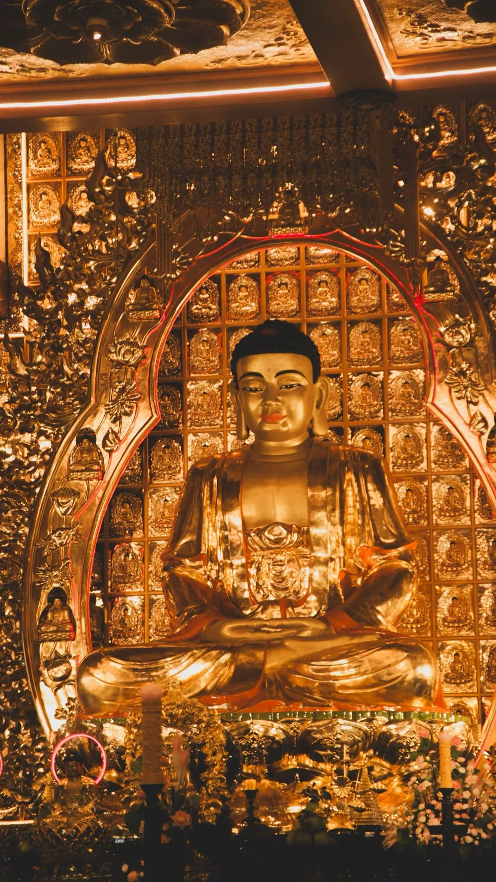 A gilded Buddha statue seated in meditation before a wall of small golden Buddha figures