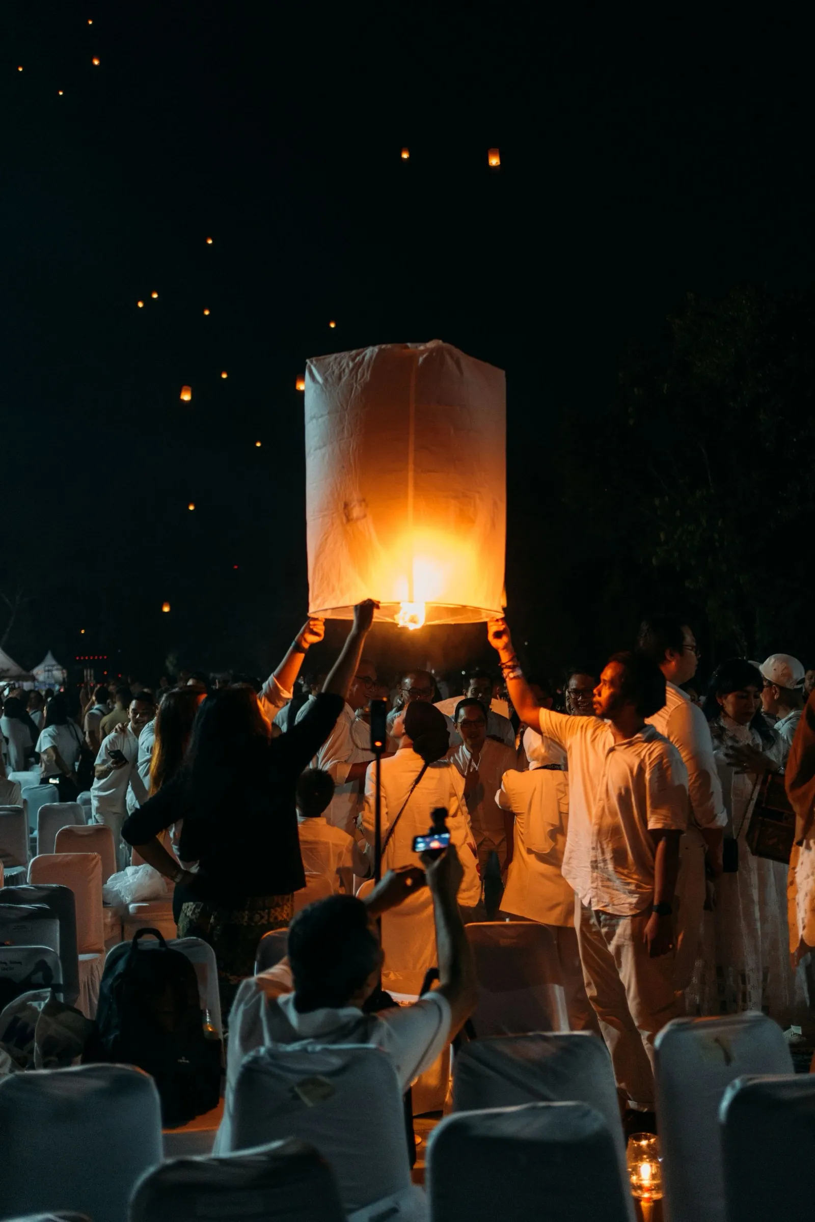 A glowing paper lantern rising into the night sky above a gathering of devotees dressed in white during Wesak